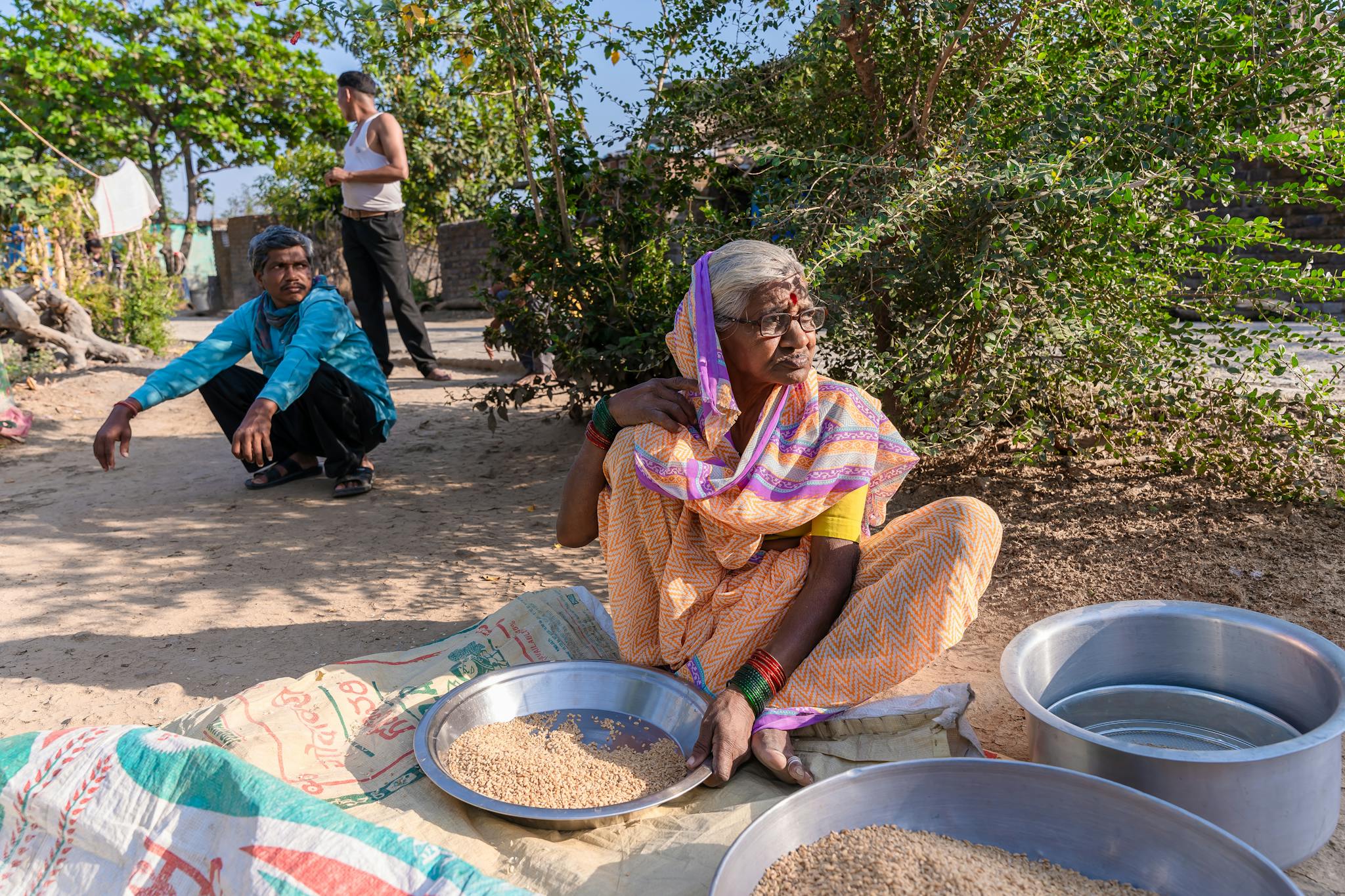 An elderly woman prepares grains outdoors in a rural Indian setting, embracing tradition.